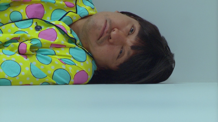 Screenshot from the film Symbol. Hitoshi Matsumoto's character is laying in a white room in a yellow and dotted pajamas, his eyes are open and glassy.